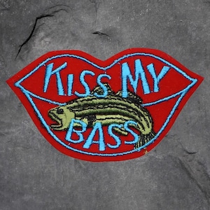 May include: A red patch in the shape of lips with the text "Kiss My Bass" in blue. A green and yellow fish is in the center of the lips. The patch has a blue border.