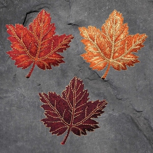 May include: Three embroidered maple leaves in different shades of red, orange, and brown. The leaves are on a dark grey background.
