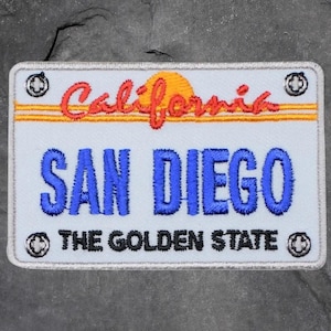 Peut inclure: Écusson brodé en forme de plaque d'immatriculation californienne. Il présente le mot "California" en rouge au-dessus de "SAN DIEGO" en bleu, et "THE GOLDEN STATE" en noir. Comprend un motif de soleil jaune et orange.