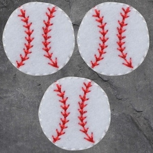 Könnte beinhalten: Drei weiße, baseballförmige Aufnäher mit roten Nahtdetails. Die Aufnäher sind in einem dreieckigen Muster auf einem dunkelgrauen Hintergrund angeordnet. Die roten Nähte ahmen die Nähte eines Baseballs nach.