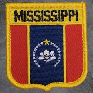 May include: Embroidered patch with the word "MISSISSIPPI" in black on a yellow border. The patch shows the state flag, with red side panels and a blue central section featuring a white flower and star design.