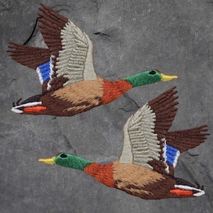 May include: Two embroidered mallard ducks in flight, one brown and one tan, with green heads and yellow beaks, against a gray slate background.
