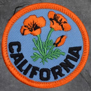 May include: Round patch featuring orange California poppies on a light blue background, with the word "CALIFORNIA" in black lettering. The patch has an orange border and green stems and leaves.