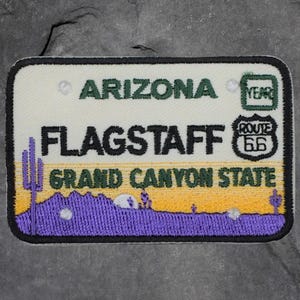 May include: Embroidered patch in the shape of an Arizona license plate. The patch features the words "ARIZONA", "FLAGSTAFF", and "GRAND CANYON STATE". A Route 66 logo is on the right. The background shows a desert landscape with purple mountains and a yellow sky.