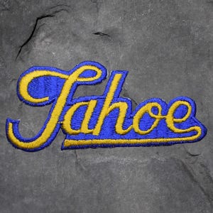 Könnte beinhalten: Gestickter Aufnäher mit dem Wort "Tahoe" in Schreibschrift. Der Text ist gelb mit blauem Rand und befindet sich auf blauem Grund. Der Aufnäher ist wahrscheinlich für eine Jacke oder eine Tasche.