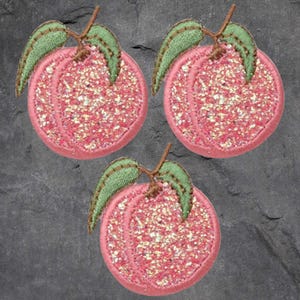 May include: Three embroidered patches in the shape of peaches. Each peach is pink with glitter and has green leaves and a brown stem. The patches are circular and have a stitched border.