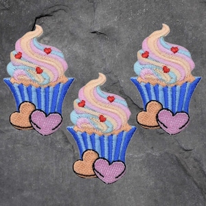 May include: Three embroidered iron-on patches of pastel pink, blue, and yellow cupcakes with red hearts and blue and white striped cupcake liners. Each cupcake has two pink hearts at the bottom.