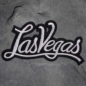 May include: Embroidered patch featuring the text "Las Vegas" in a cursive font. The lettering is white, outlined in black, and set against a black background. The patch is likely for clothing or accessories.