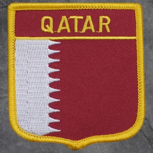 May include: A maroon and white patch with the word "QATAR" in yellow. The patch has a yellow border and a white serrated edge on the left side, resembling a flag.