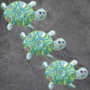 May include: Three embroidered turtle patches. Each turtle has a light green body with blue and green stitching, and silver sequins. The turtles have smiling faces and light green flippers.