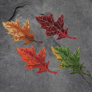 May include: Four embroidered oak leaves in autumnal shades of orange, red, burgundy, and green. Each leaf has detailed stitching with gold thread accents, set against a dark grey, textured background. The leaves are arranged in a circular pattern.