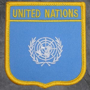 Peut inclure: Un écusson bleu avec l'inscription "UNITED NATIONS" en lettres dorées. Il présente un emblème blanc du planisphère entouré de branches d'olivier. La bordure est jaune.