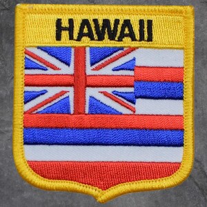 May include: Embroidered patch featuring the word "HAWAII" in black letters above a flag design. The flag incorporates the Union Jack in the upper left corner, with red, white, and blue stripes below. The patch has a yellow border.