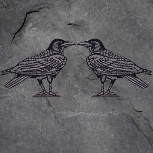 Könnte beinhalten: Zwei gestickte Raben-Patches, die sich gegenüberstehen, mit detaillierten Federstrukturen in Grau- und Schwarztönen. Die Patches sind vor einem strukturierten, dunkelgrauen Hintergrund platziert, was einen auffälligen Kontrast erzeugt.