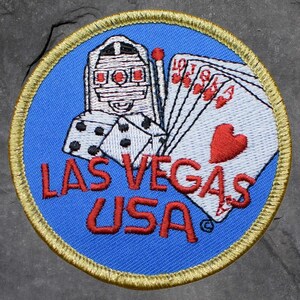 Peut inclure: Écusson brodé avec un fond bleu et une bordure dorée. Le patch représente une machine à sous, des dés, des cartes à jouer et le texte "LAS VEGAS USA" en rouge.