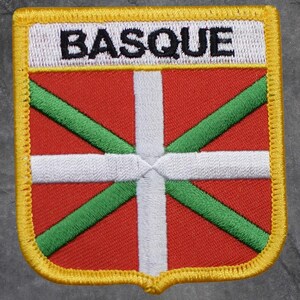 May include: Embroidered patch featuring the Basque flag design. The patch has a red background with a white cross and green diagonal lines. The word "BASQUE" is embroidered in black on a white banner at the top. The patch is bordered with yellow stitching.