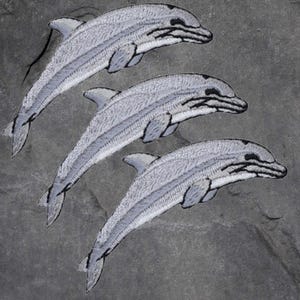 May include: Three grey embroidered dolphin patches. The dolphins are facing right and are in a leaping position.