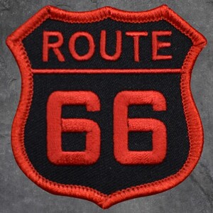 Könnte beinhalten: Ein Route 66-Aufnäher mit schwarzem Hintergrund und roten Stickdetails. Der Aufnäher zeigt die Worte "ROUTE" über der Nummer "66" in Schildform mit rotem Rand.