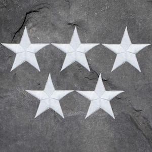 May include: Five white star-shaped patches arranged in a pattern on a dark gray, textured surface. The stars have a slightly raised, embroidered appearance, with visible stitching details. The patches are evenly spaced and appear to be made of fabric.