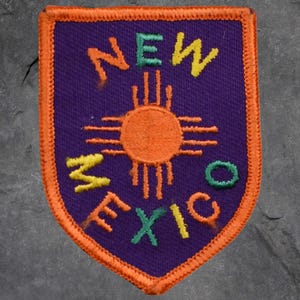 May include: Embroidered patch with the text "NEW MEXICO" in yellow, green, and orange. The patch has a purple background, an orange sun symbol, and an orange border.