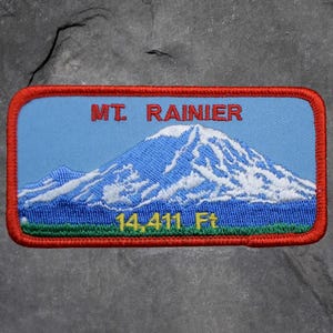 May include: Embroidered patch featuring Mt. Rainier with a blue sky background. The mountain is depicted in white and blue, with the text "MT. RAINIER" above and "14,411 Ft" below, all within a red border.