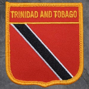 May include: A rectangular patch featuring the flag of Trinidad and Tobago. The patch has a red background with a black diagonal stripe edged in white. The words "TRINIDAD AND TOBAGO" are embroidered in gold across the top, with a gold border.