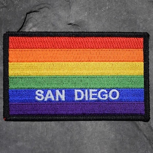 May include: Rectangular embroidered patch featuring the rainbow flag design with the text "SAN DIEGO" in white. The patch has a black border and is set against a textured gray background.