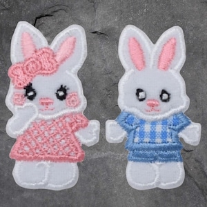 May include: Two white felt bunny appliques, one wearing a pink dress with white polka dots and the other wearing a blue and white checked shirt.
