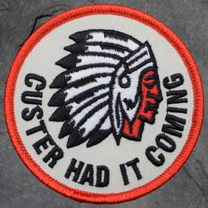 Peut inclure: Écusson brodé circulaire avec un motif de chef amérindien. L'écusson présente un fond crème, une bordure rouge et le texte "Custer Had It Coming" en noir. Le visage du chef est rouge, avec une coiffe blanche et noire.