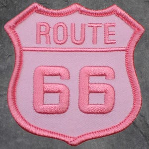 May include: A pink Route 66 patch with embroidered text and border. The shield-shaped patch features the words "ROUTE" above the number "66". The design is in shades of pink, with a darker pink border.