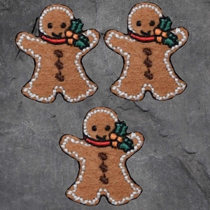 May include: Three gingerbread man ornaments. Each ornament is brown with white icing trim, a red scarf, and a sprig of green and orange holly. The gingerbread men have a smiling face and three brown button details.