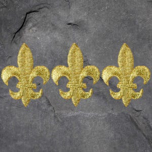 May include: Three gold fleur-de-lis patches, embroidered with a textured finish. The patches are arranged horizontally against a dark gray, textured background.