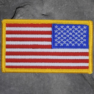 May include: Embroidered patch featuring the American flag. The patch has a yellow border, red and white stripes, and a blue square with white stars. The patch is rectangular in shape.