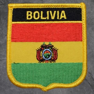May include: Embroidered patch featuring the Bolivian flag. The patch has a black top section with the word "BOLIVIA" in yellow. Below are horizontal stripes of red, yellow, and green. The yellow stripe includes a coat of arms. The patch is bordered with yellow.