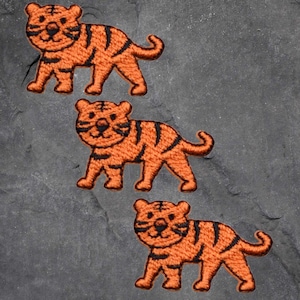 May include: Three orange tiger patches with black stripes are arranged diagonally on a dark gray surface. Each patch features a smiling tiger design, with detailed embroidery creating a textured appearance. The patches are likely for decorative purposes.