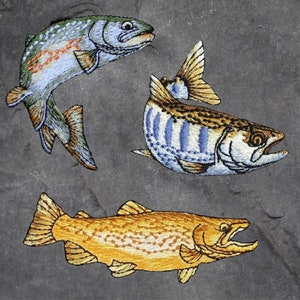 May include: Embroidered patches depicting three fish. One is light blue and green with orange details, another has blue stripes, and the third is golden yellow. The patches are set against a grey background.