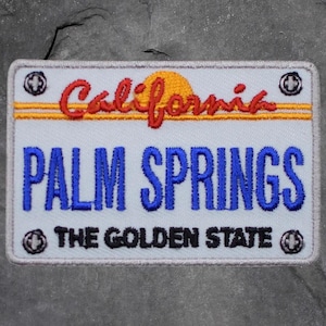 May include: Embroidered patch in the shape of a California license plate. The patch features the words "California" in red script, "PALM SPRINGS" in blue, and "THE GOLDEN STATE" in black. A yellow sun and border complete the design.