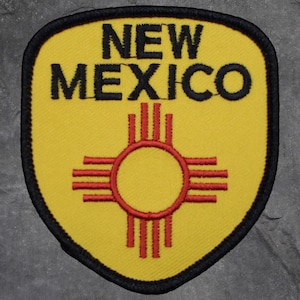 以下が含まれることがあります： 黒い文字で「NEW MEXICO」と書かれた黄色のパッチ。パッチの中央には赤と黄色のジアシンボルがあり、黒い縁取りがあります。