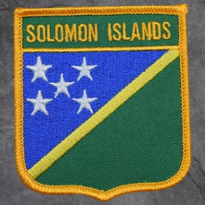 May include: Embroidered patch featuring the text "SOLOMON ISLANDS" in gold on a green background. The patch has a blue field with five white stars and a green and yellow diagonal stripe. The border is gold.