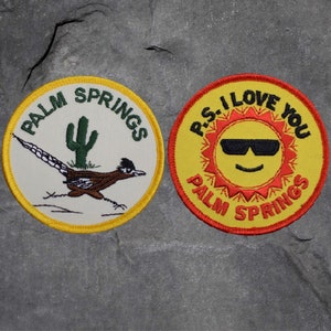 May include: Two round embroidered patches. One features a roadrunner and cactus with "PALM SPRINGS" text. The other has a smiling sun wearing sunglasses, with "P.S. I LOVE YOU" and "PALM SPRINGS" text. Both patches have yellow borders.