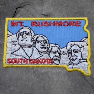 May include: Embroidered patch depicting Mount Rushmore, with the text "MT. RUSHMORE SOUTH DAKOTA." The patch is shaped like South Dakota, with a blue background and a yellow border. The presidential faces are white.
