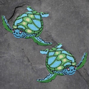以下が含まれることがあります： 緑と青の貝殻を持つ、刺繍されたウミガメのパッチが2つ。カメは異なる方向を向いており、黒い目があります。