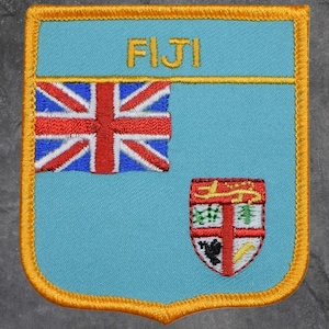 May include: Embroidered patch featuring the Fiji flag and coat of arms. The patch has a light blue background with a gold border. The flag includes the Union Jack and the coat of arms has a red cross and other symbols. The word "FIJI" is at the top.
