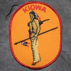 May include: Embroidered patch with an orange background and a red border. The patch features the word "KIOWA" in red at the top. A Native American figure is in the centre, holding a spear. The figure is wearing a headdress and traditional clothing.