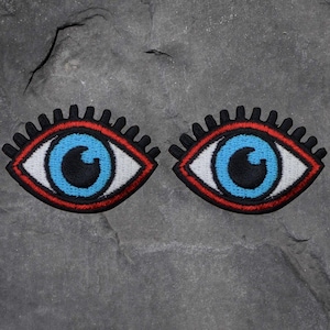 May include: A pair of embroidered eye patches. Each patch has a blue iris with a black pupil, a white sclera, a red outline, and black eyelashes. The patches are on a grey, textured background.