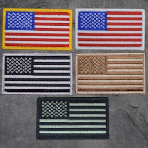 以下が含まれることがあります： 異なる色のバリエーションで刺繍されたアメリカ国旗のパッチが4つ。パッチはグレーのテクスチャのある背景の上にあります。パッチは黒と白、茶色とベージュ、赤、白、青、緑と黒です。