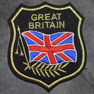 May include: A black embroidered patch with the words "GREAT BRITAIN" in gold lettering. The patch features the Union Jack flag in red, white, and blue, and is bordered with gold stitching.