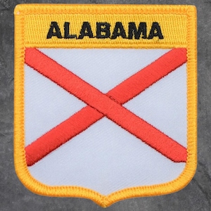 May include: Embroidered patch with the word "ALABAMA" in black on a yellow background. The patch has a white centre with a red cross, and a yellow border. The patch is shield-shaped.