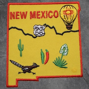 May include: Embroidered patch in the shape of New Mexico, featuring the state name in red, Route 66, a hot air balloon, a roadrunner, cactus, and chili peppers. The patch has a yellow background with a red border.