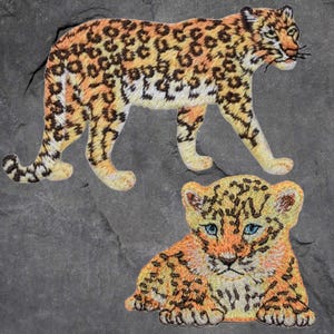 May include: Two embroidered patches of a leopard, one adult and one cub. The adult leopard is walking and has a brown, orange, and white spotted pattern. The cub is lying down and has a similar spotted pattern.